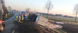Zdjęcie przedstawia samochód ciężarowy leżący na prawym boku w przydrożnym rowie, z którego częściowo wysypał się ładunek w postaci płyt drewnopodobnych. Ziemia częściowo znajduje się na jezdni. Na zdjęciu widać również samochód strażacki i strażaków. 