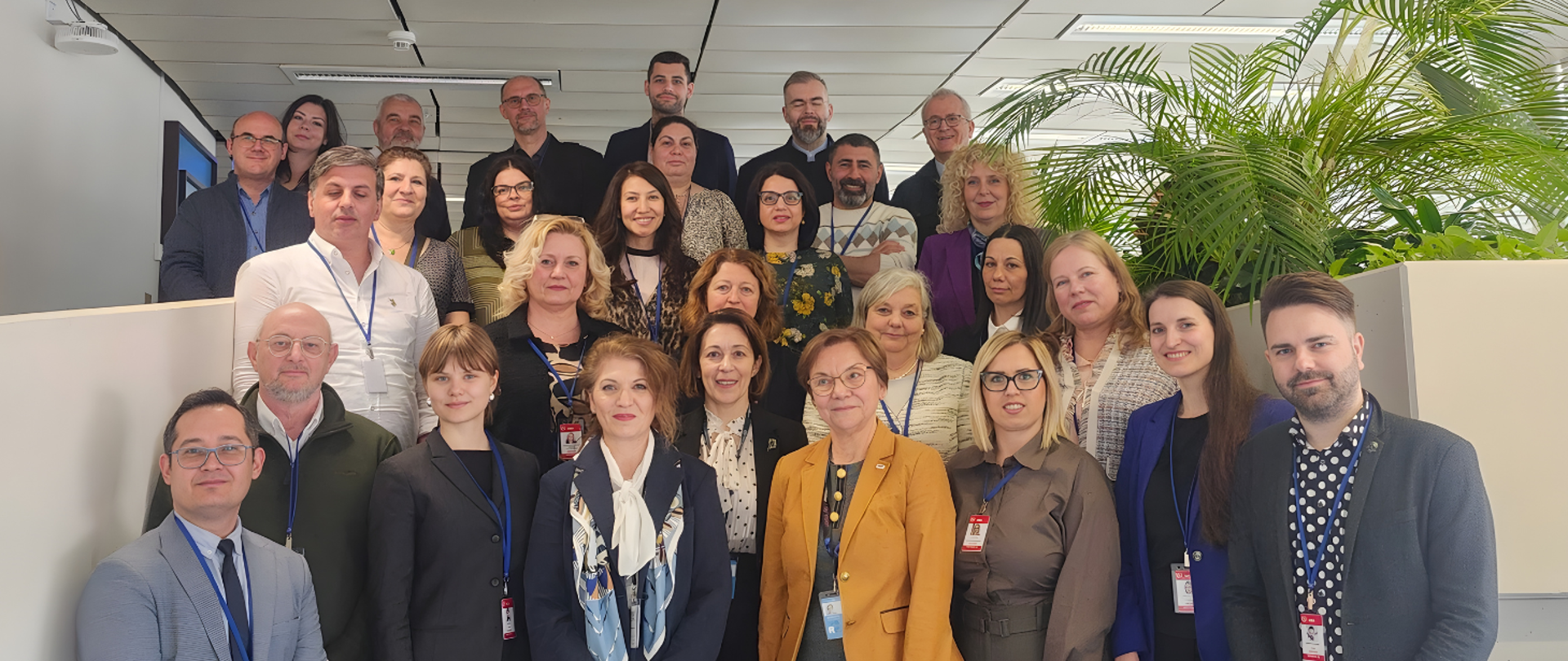 Zdjęcie grupowe około trzydziestu pracowników pozujących w biurze. Osoby stoją w kilku rzędach przy jasnych boksach biurowych, większość ma identyfikatory na niebieskich smyczach i ubrana jest w stroje biznesowe lub smart casual. Po prawej stronie widać duże zielone rośliny, a w tle sufit z oświetleniem biurowym.
