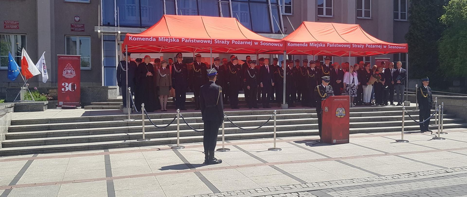 Na zdjęciu Komendant Miejski PSP w Sosnowcu podczas przemówienia. W tle trybuna honorowa z przybyłymi gośćmi.