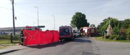 Potrącenie rowerzysty przez samochód dostawczy, zdjęcie zrobione z dala, widać pojazdy służb, oraz czerwony parawan zabezpieczający teren działań ratowniczych