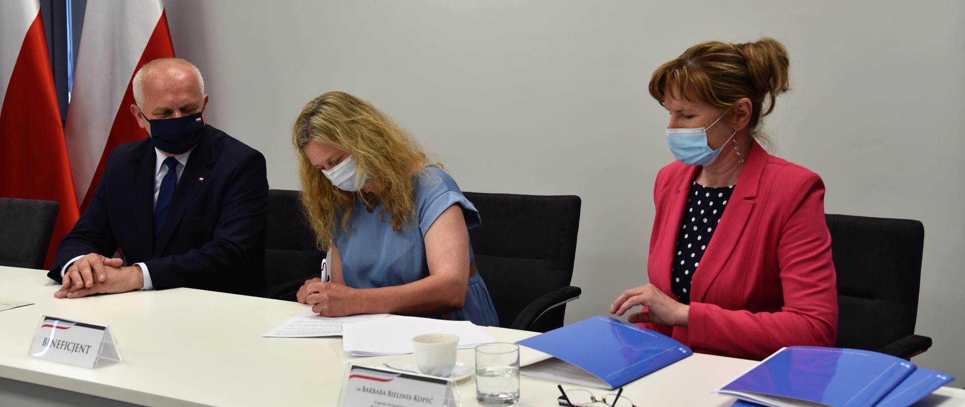 Przy białym stole, na tle białej ściany siedzi wojewoda, beneficjentka i konserwator zabytków podpisując umowę. 