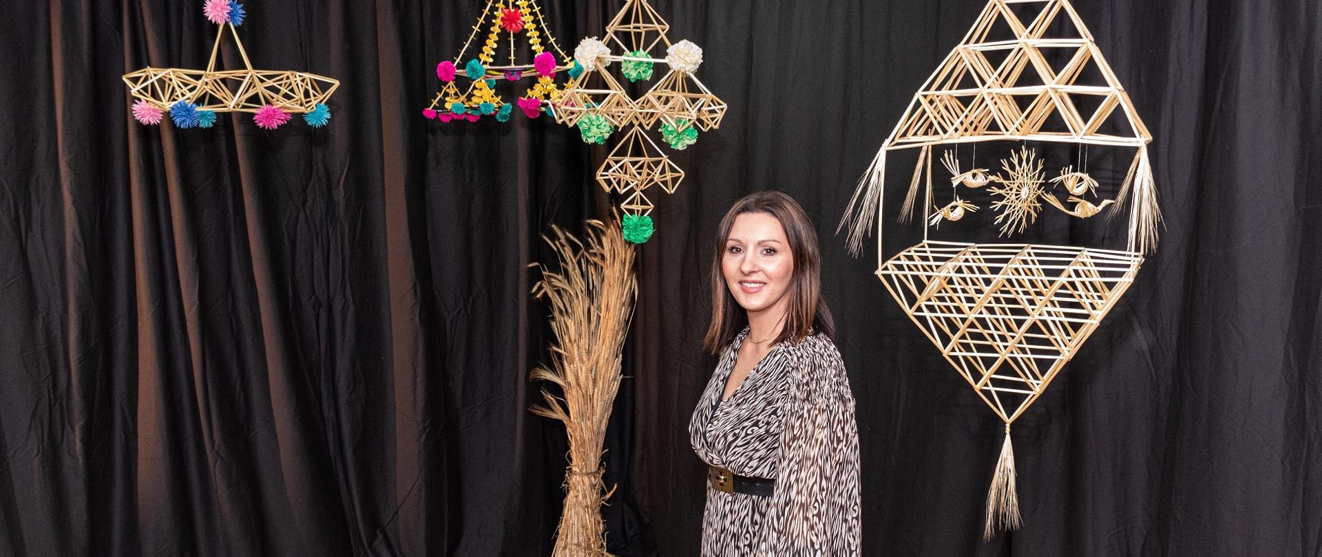  Na zdjęciu autorka pracy dyplomowej Elżbieta Florek. Stoi na czarnym tle gdzie przy suficie są powieszone jej prace czyli ozdoby, tzw. pająki ze słomy. 