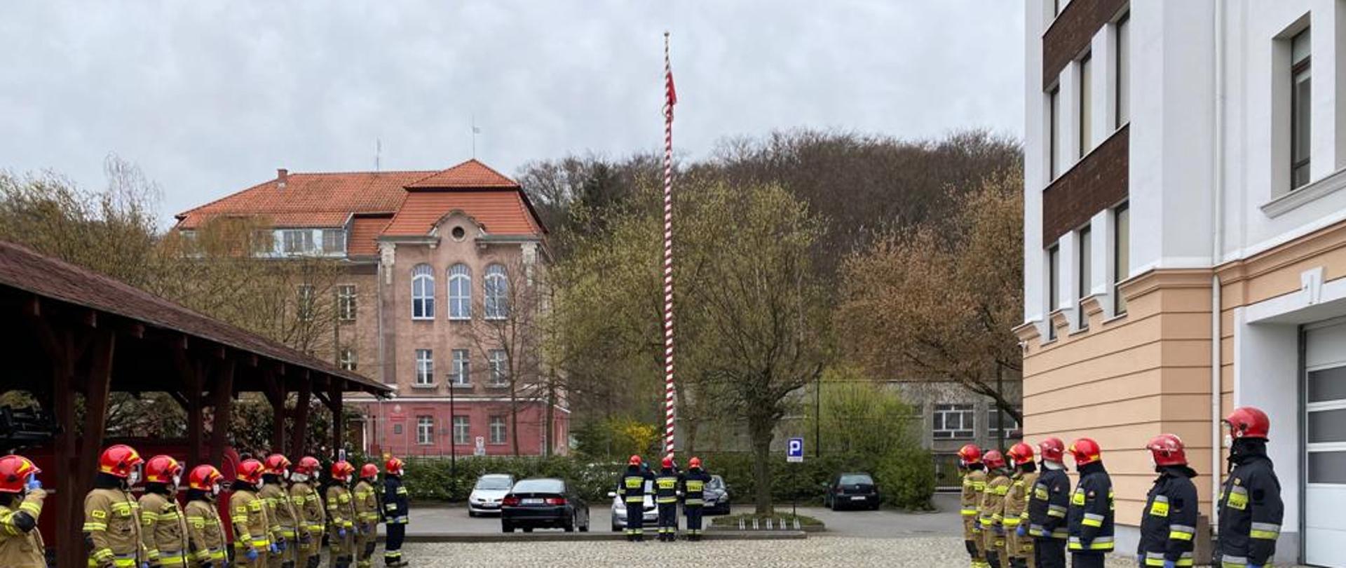 Strażacy stoją w dwóch rzędach na przeciwko siebie, uroczyście podnosząc flagę Rzeczypospolitej Polskiej na maszt. 