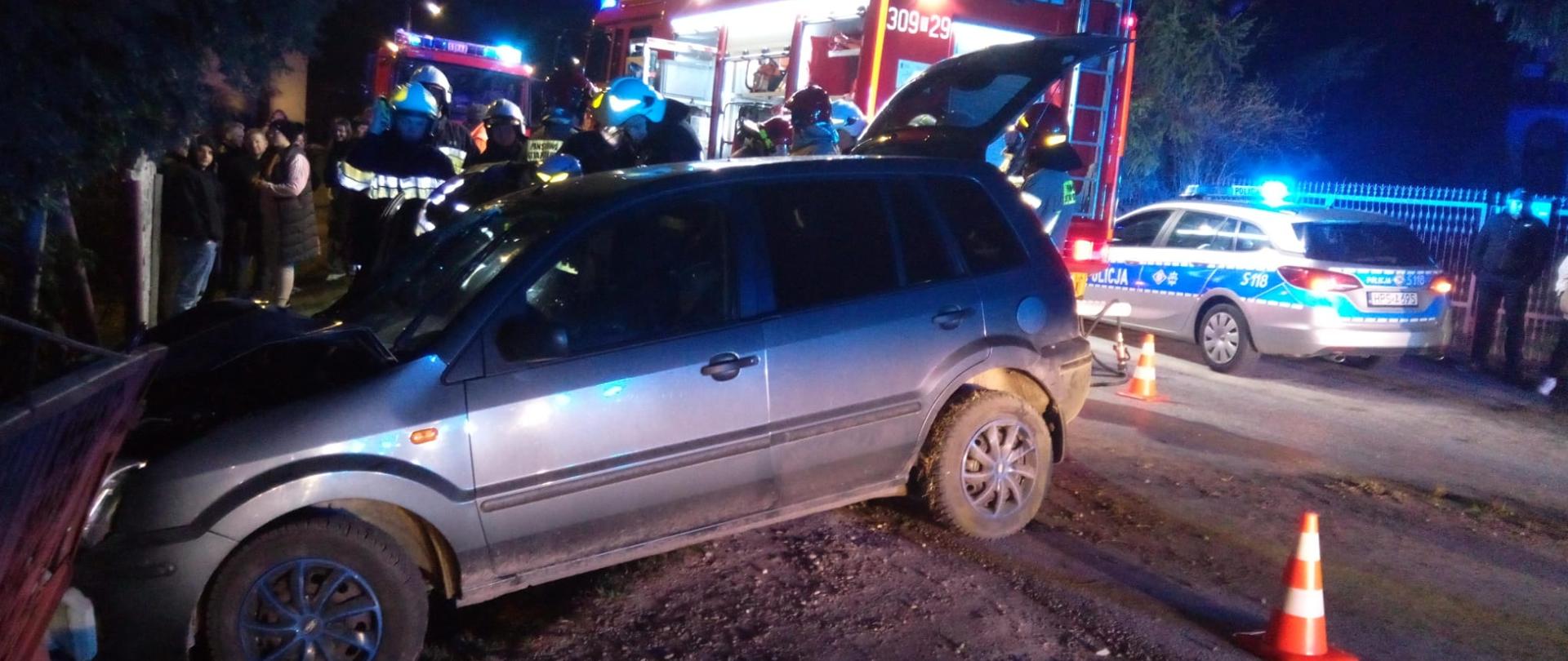 Zdjęcie przedstawia samochód osobowy, który wypadł z drogi i uderzył w ogrodzenie. Przed autem widać pachołek oznakowania terenu. Za pojazdem widać wóz strażacki, strażaków a z prawej strony radiowóz policyjny.