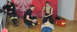 Egzamin recertyfikacyjny. Świetlica Komendy Powiatowej PSP w Rawiczu. Na tle ścianki promującej komendę dwóch egzaminowanych strażaków ratowników OSP udziela kwalifikowanej pierwszej pomocy "poszkodowanemu" manekinowi. Jeden z nich klęcząc z boku obsługuje AED, drugi, trzymając w ręce worek AMBU oczekuje na wskazania defibrylatora. Z prawej strony stoi torba medyczna. Przy stole siedzi członek komisji egzaminacyjnej.