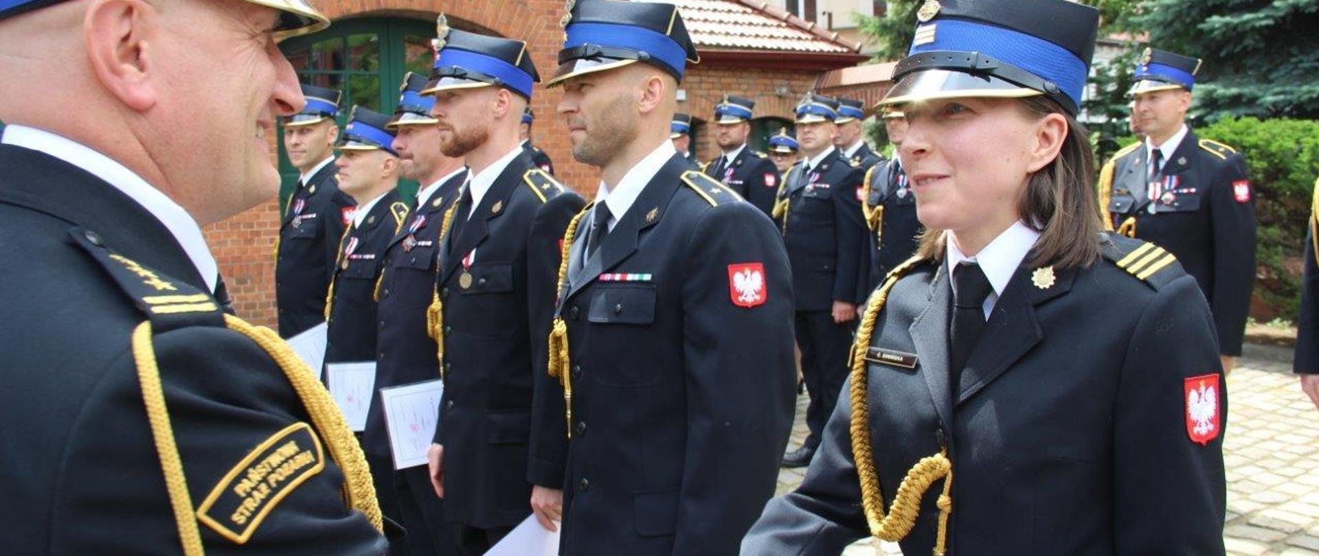 Przełożony składa gratulacje funkcjonariuszce, która stoi w szeregu innych strażaków. Wszyscy w mundurach galowych. W tle budynek komendy z zegarem na elewacji.