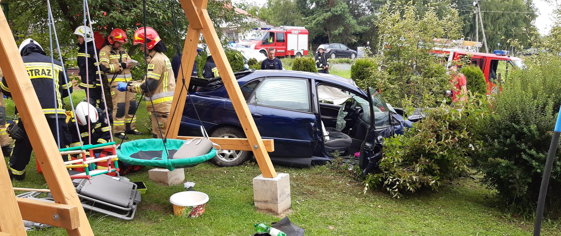Na zdjęciu widoczny uszkodzony samochód na posesji. Przed samochodem stoi huśtawka dziecięca. Przy samochodzie znajdują się strażacy. W oddali na drugim planie widoczni policjanci i samochód pożarniczy. 