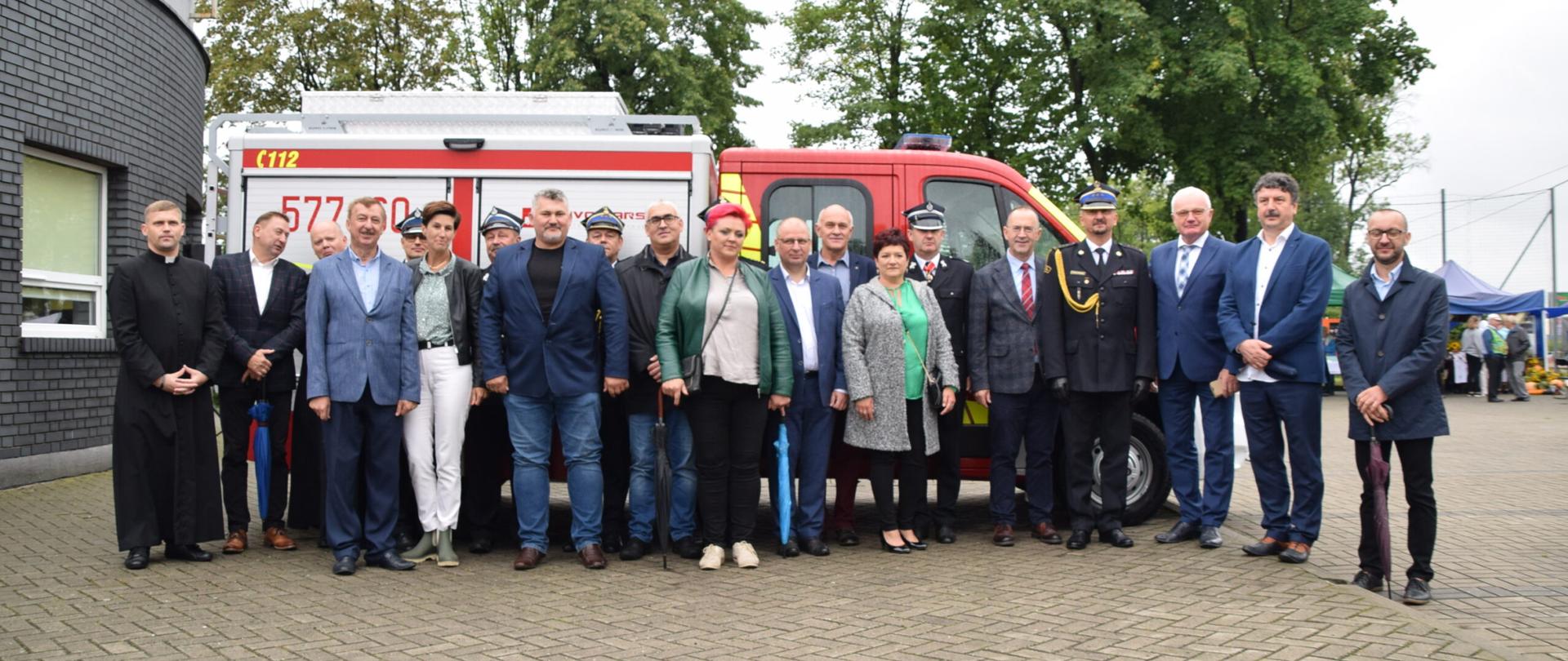 kilkanaście osób stojących przed samochodem gaśniczy, pośród osób kilku strażaków w mundurach wyjściowych w tle drzewa