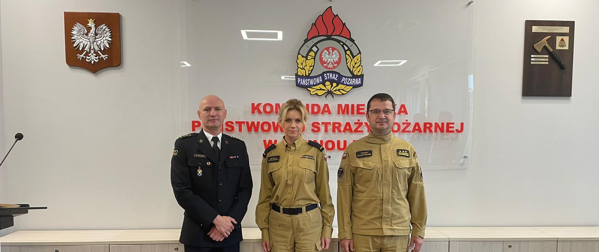 Pamiątkowe zdjęcie. Funkcjonariusze stoją w sali konferencyjnej na tle napisu "Komenda Miejska Państwowej Straży Pożarnej". Od lewej Komendant Miejski, następnie przenoszony funkcjonariusz oraz Zastępca Komendanta Miejskiego.
