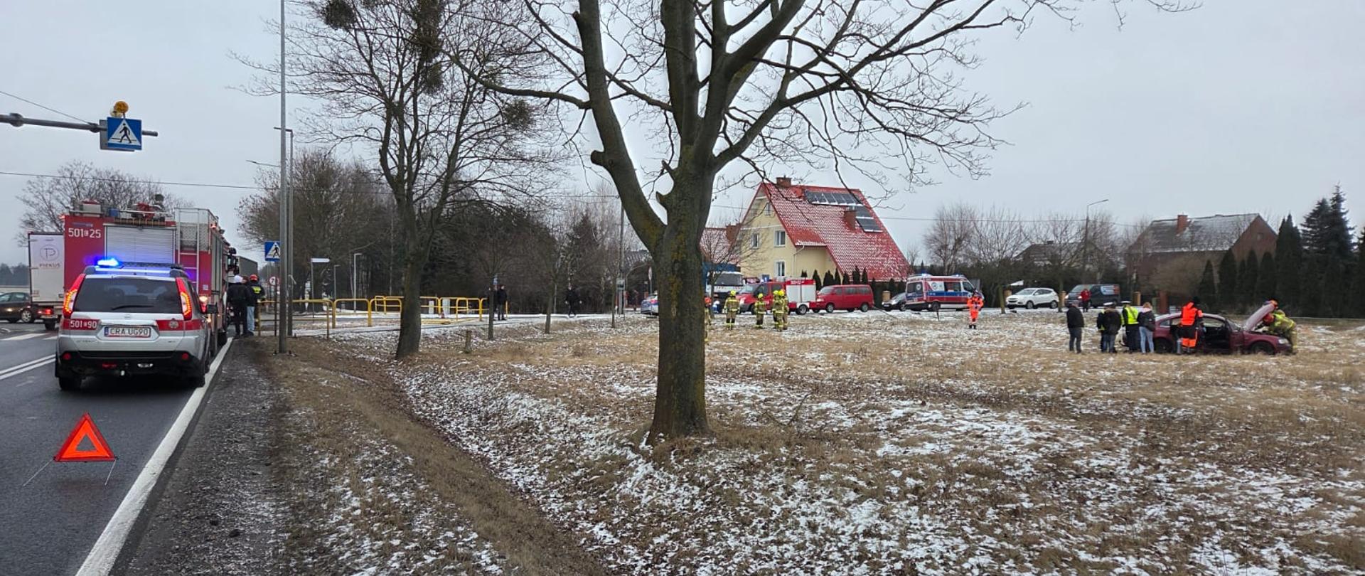 Na zaśnieżonym polu stoi uszkodzony samochód osobowy z otwartą maską, w pobliżu znajdują się funkcjonariusze Policji oraz strażacy prowadzący działania zabezpieczające.