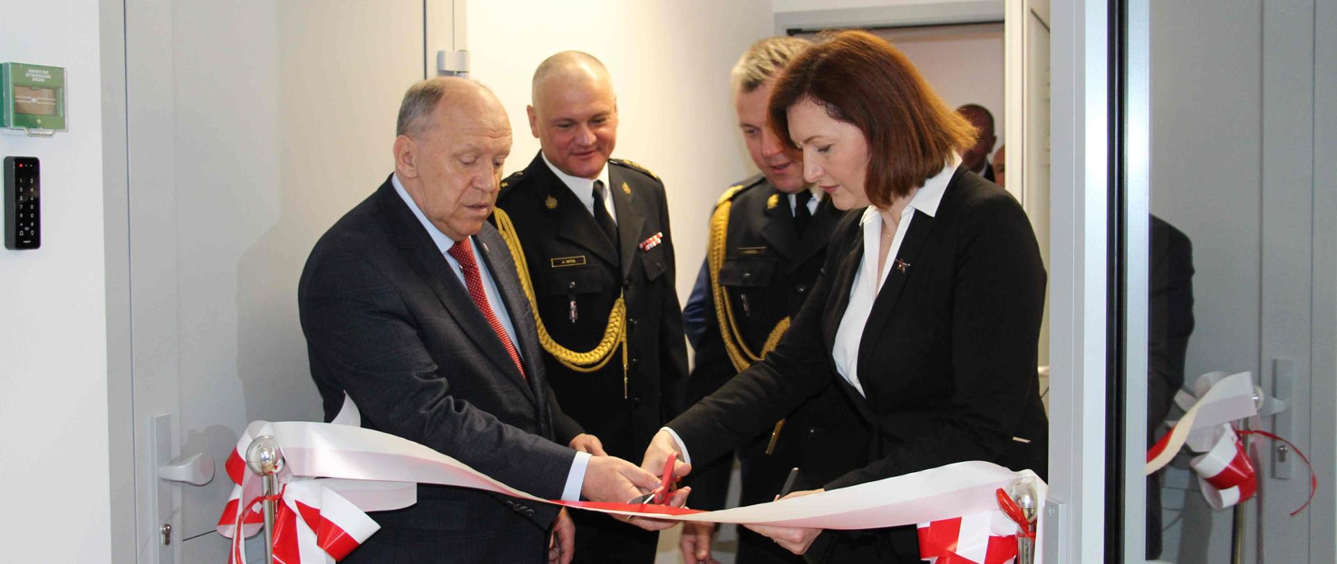 W wąskim pomieszczeniu stoi mężczyzna w garniturze, kobieta oraz dwóch strażaków. W przejściu pomiędzy drzwiami przewieszona biało czerwona wstęga, którą wspólnie przecinają nożyczkami mężczyzna i kobieta