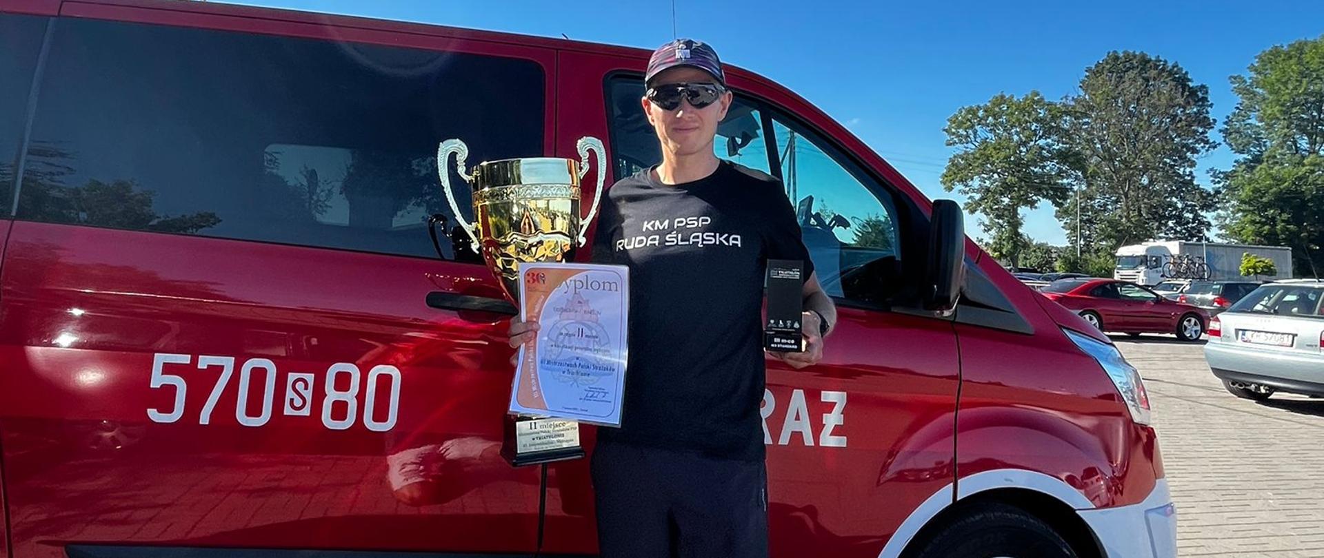 Zdjęcie przedstawia strażaka w tle samochodu pożarniczego po zdobyciu 2 miejsca na mistrzostwach polski w triathlonie.