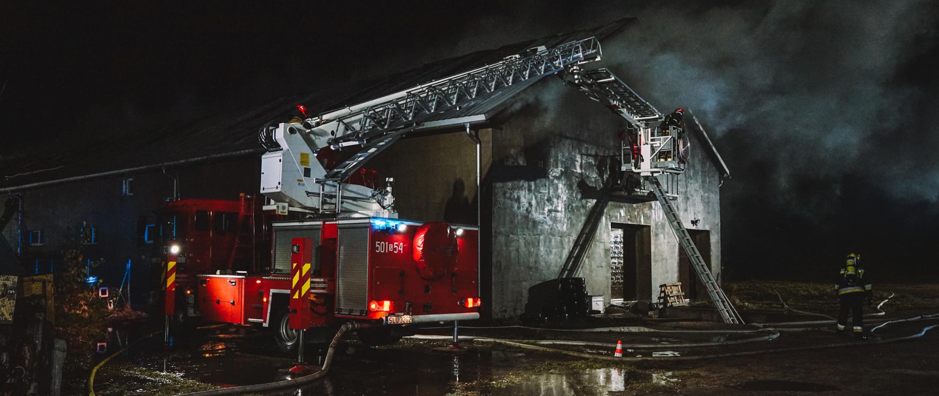 Na zdjęciu widać podnośnik hydrauliczny z JRG Lubliniec z rozstawionym koszem. W koszu strażak PSP w ubraniu specjalnym, hełmie bojowym i aparatem ochrony układu oddechowego w bezpośrednim sąsiedztwie budynek objęty pożarem, spod dachu widać kłęby gęstego dymu