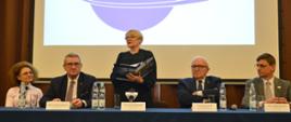 Przy nakrytym niebieskim materiałem stole siedzą cztery osoby, pośrodku stoi kobieta w czarnym ubraniu i mówi trzymając w rekach otwartą książkę, za nimi na wielkim ekranie napis CBK PAN.