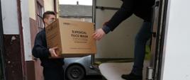 Strażak JRG Wadowice stoi za samochodem dostawczym i trzyma w rękach paczkę z maseczkami ochronnymi, przekazując ją osobie, która stoi na skrzyni ładunkowej auta dostawczego.