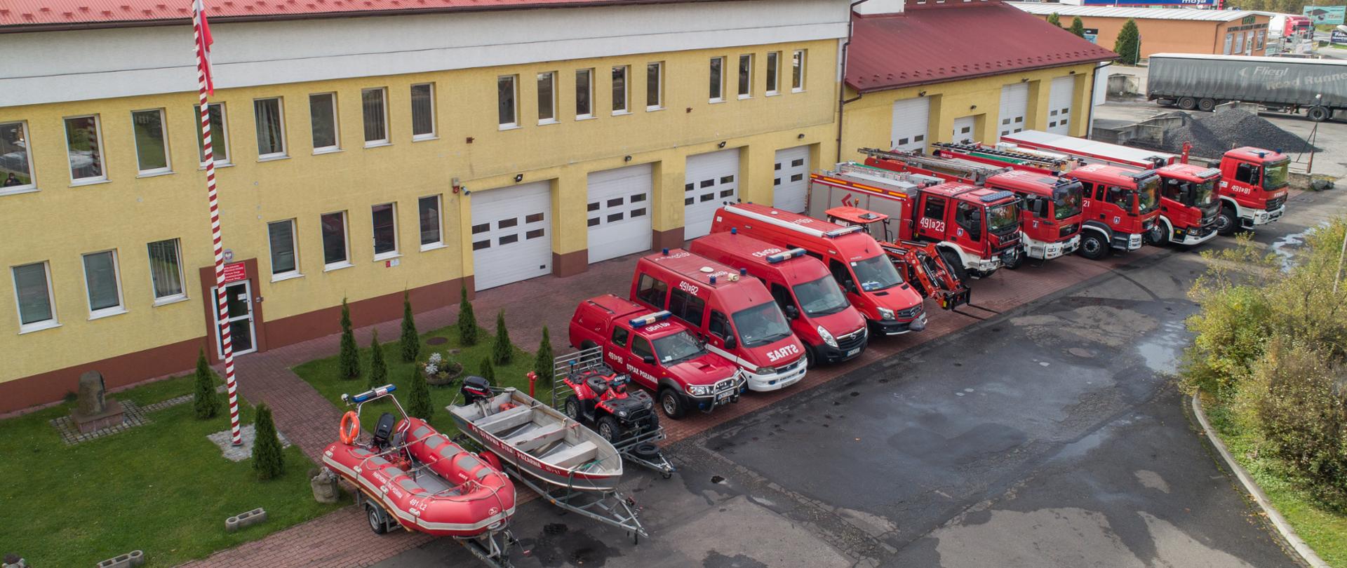 Zdjęcie budynku Jednostki ratowniczo-gaśniczej w Nowym Targu. Na tle budynku powiewa flaga państwowa. Przed garażami zostały wystawione pojazdy pożarnicze, którymi dysponuje jednostka. Pojazdy są w kolorze czerwonym, i są to trzy samochody ratowniczo-gaśnicze, jeden samochód ratownictwa chemicznego, jeden samochód kontener i dwa busy, samochód ratownictwa wodnego, samochód terenowy, quad, traktor, łódź i ponton. Wokół budynku znajdują się zagospodarowane tereny zielone: drzewa i krzewy. Na drugim planie zdjęcia znajduje się skład materiałów opałowych.Zdjęcie zostało wykonane z drona.