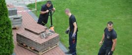 Remont chodnika. Teren przed budynkiem Komendy Powiatowej PSP w Rawiczu. Trzech strażaków, którzy wykonują prace remontowe. Jeden z nich operuje wózkiem widłowym, na którym na drewnianej palecie ułożona jest kostka pochodząca z rozbiórki chodnika. Dwóch pozostałych stoi obok. Wokół chodnika - zieleń.