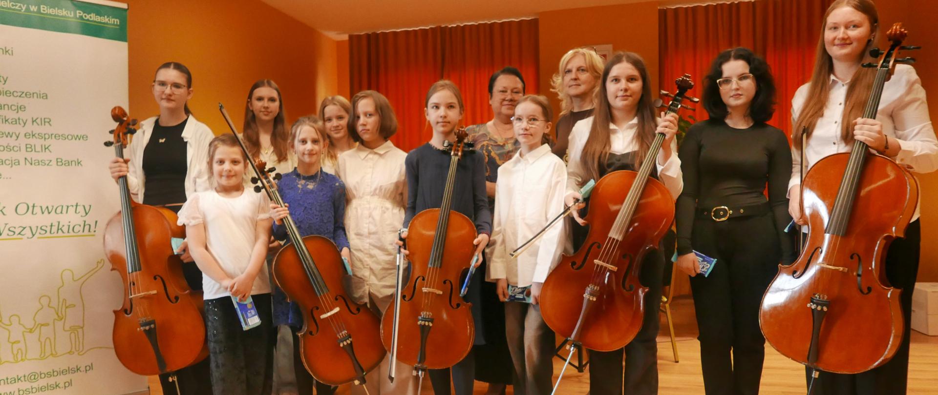Na zdjęciu widzimy same dziewczyny wchodzące w skład klasy wiolonczeli. Widzimy też nauczycielkę przedmiotu i akompaniatorkę. Wszystkie dziewczęta będące na pierwszym planie trzymają wiolonczele.