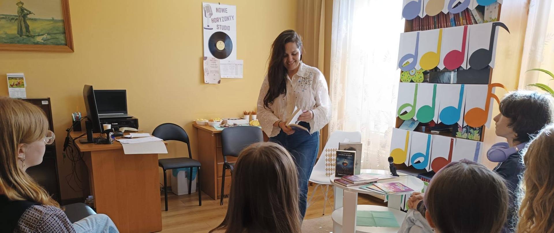 Kobieta czyta książkę grupie młodych osób siedzących w kręgu w przytulnej sali. W tle widać dekoracje z nutami muzycznymi, stolik z książkami oraz regał z książkami.