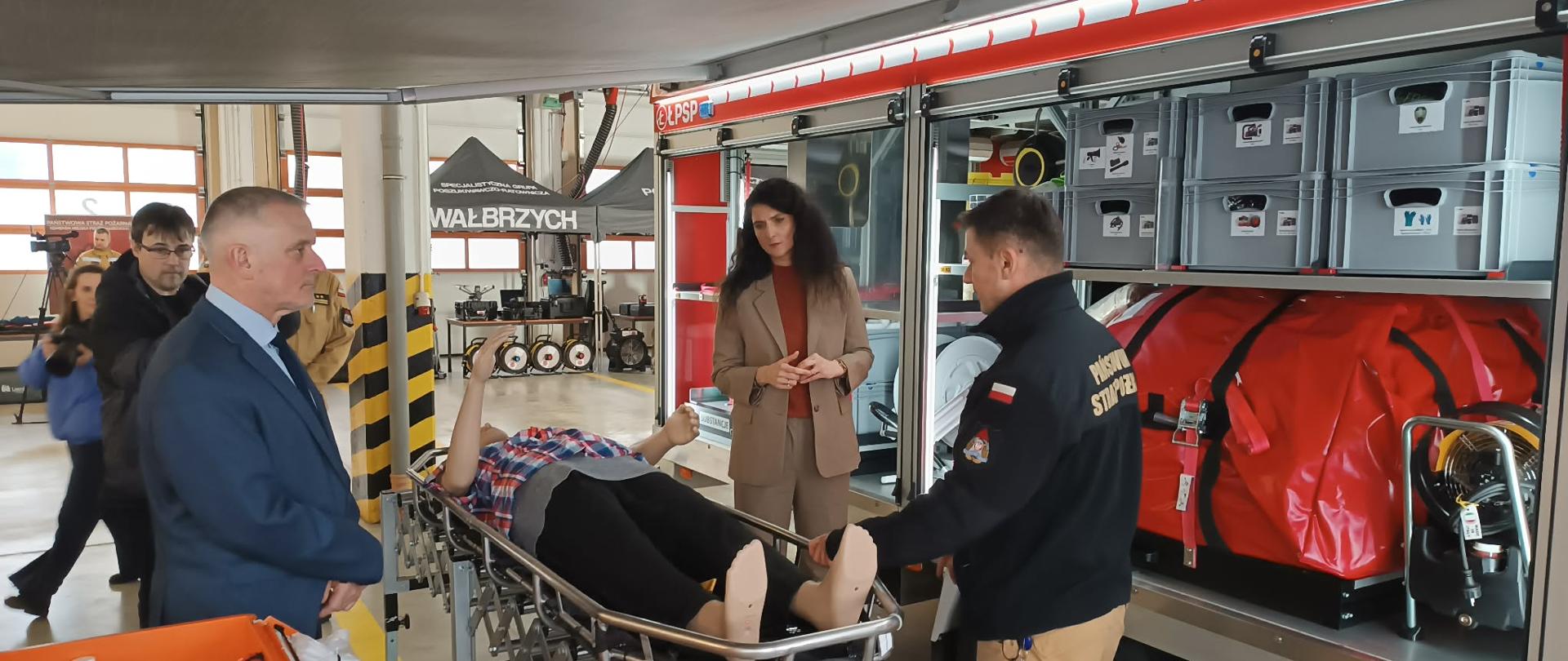 Grupa osób stoi obok rozłożonych noszy podbierakowych zamontowanych na szynach przyczepy logistycznej. Strażak objaśnia sposób transportu poszkodowanego (fantoma). Scena pokazuje praktyczne zastosowanie modułowej zabudowy przyczepy w ratownictwie.