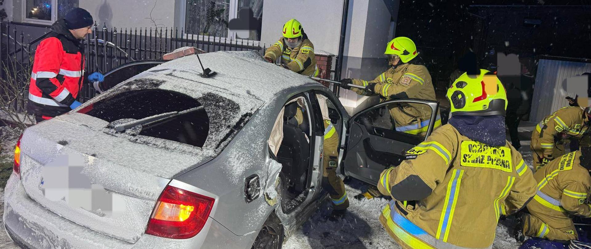 Na zdjęciu widać rozbity srebrny samochód osobowy. Pojazd nie ma tylnych prawych drzwi a przednie są otwarte. Dookoła pojazdu widać strażaków w żółtych strojach. Z lewej strony widać również ratownika medycznego. W tle widać dom. Jest ciemno, ale miejsce akcji jest oświetlone. Pada śnieg.