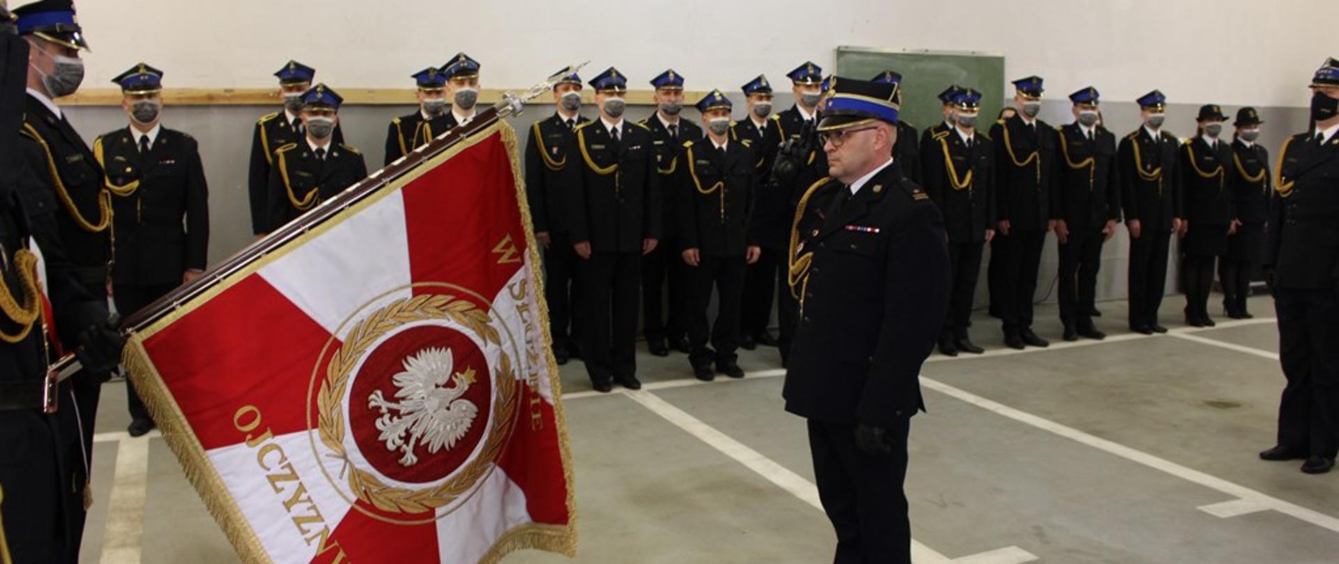 pod scianą w dwuszeregu stoją strażacy, widać pochylony sztanadar oraz strażaka stojacego na baczność przed sztandarem