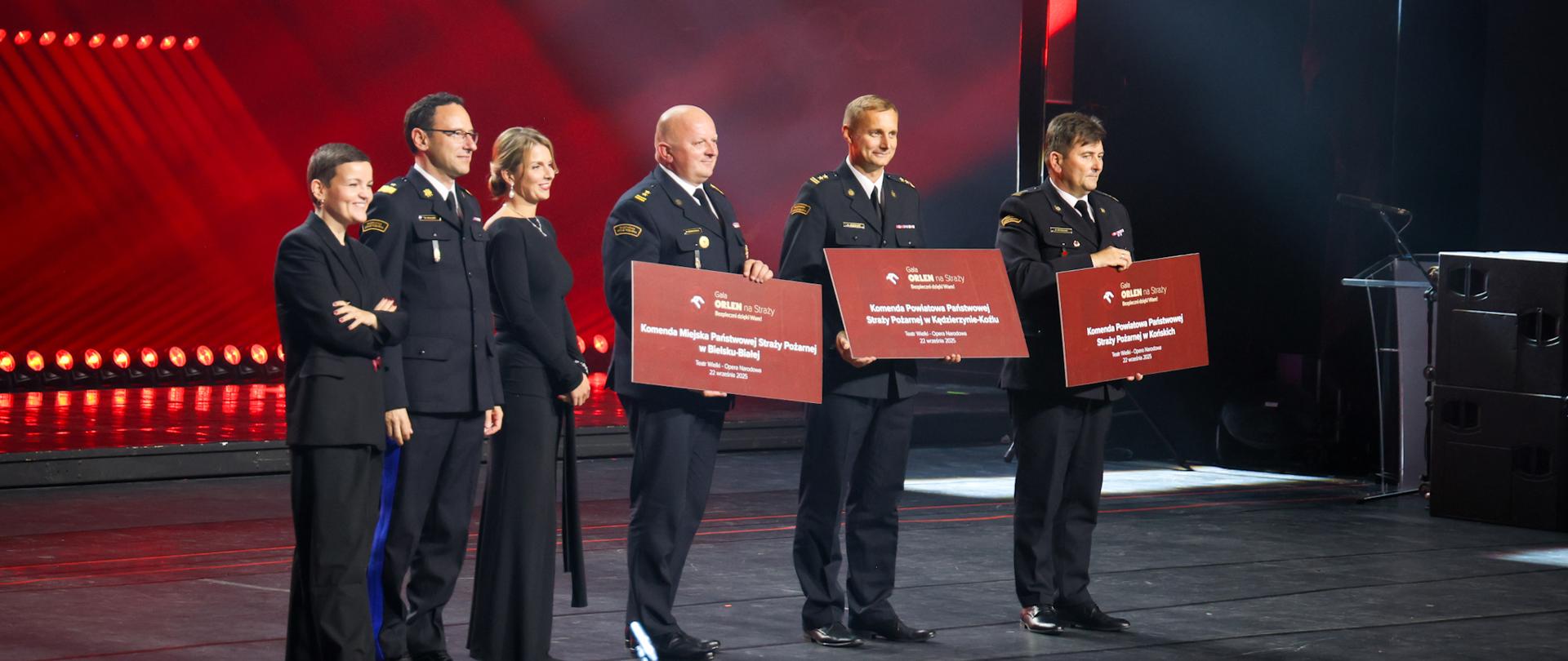 Grupa sześciu osób na scenie, w tym trzech funkcjonariuszy PSP w galowym umundurowaniu, trzymających tablice z nazwami komend PSP (Bielsko-Biała, Kędzierzyn-Koźle, Końskie); reszta to osoby cywilne w strojach wizytowych. Tło podświetlone czerwonym światłem sceny.