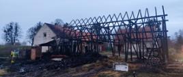Spalona konstrukcja drewnianej stodoły wraz z budynkami gospodarstwa w tle.
