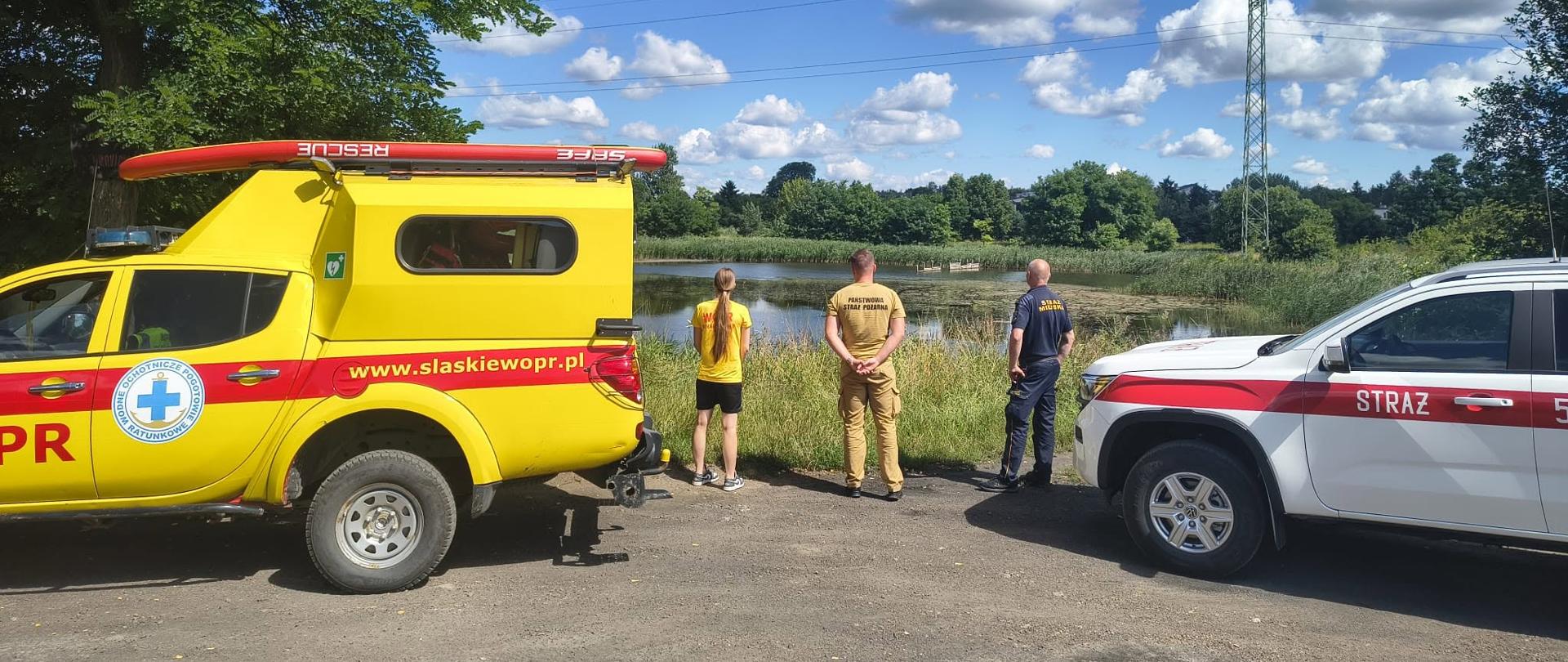 Na zdjęciu przedstawiono działania służb nad akwenem wodnym. Na pierwszym planie widoczne są pojazdy Ochotniczego Wodnego Pogotowia Ratunkowego oraz Państwowej Straży Pożarnej. Przedstawiciele służb obserwują akwen i koordynują prowadzone działania związane z poszukiwaniem osoby lub rozpoznaniem sytuacji na wodzie.