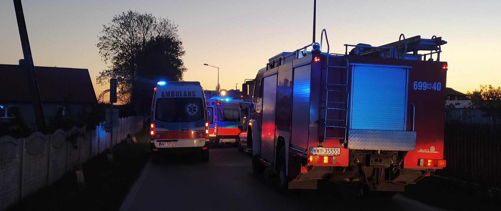 Dwie karetki pogotowia oraz pojazd pożarniczy stojące na ulicy, przy której miał miejsce pożar domu.