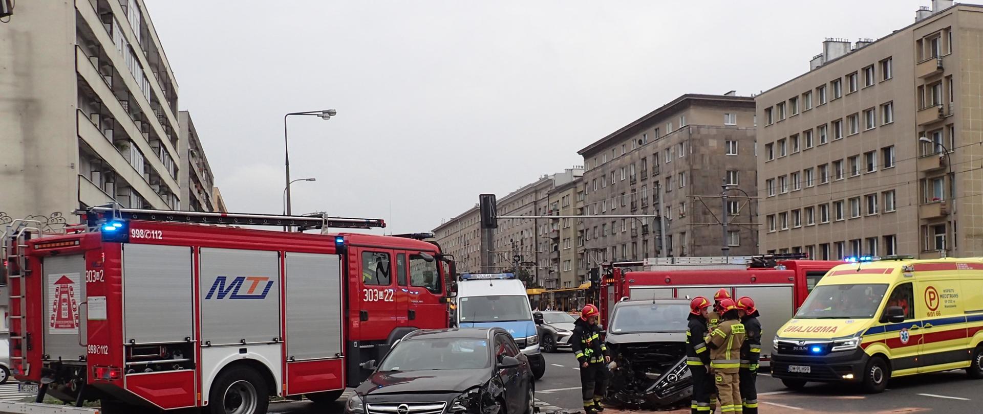 Na zdjęciu widać dwa rozbite samochody na środku skrzyżowania, w tle widoczne są dwa samochody strażackie oraz karetkę pogotowia. Przy rozbitych samochodach pracują strażacy.
