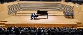 Mężczyzna gra na fortepianie na estradzie sali koncertowej, od tyłu widoczna widownia zapełniona publicznością.