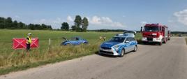 Zdjęcie przedstawia rozbite auto po dachowaniu na łące oraz wóz bojowy straży pożarnej i radiowóz policji