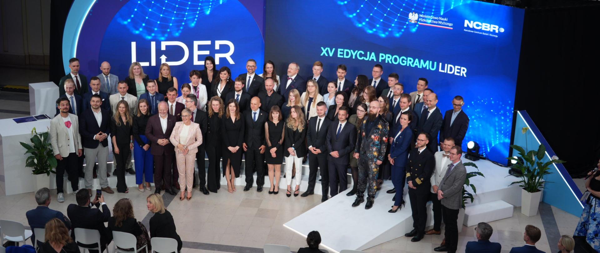 Zdjęcie od tyłu sali z góry, na sali na podwyższeniu z przodu pod wielkim niebieskim banerem z napisem LIDER - XV edycja Programu LIDER stoi grupa kilkudziesięciu osób.