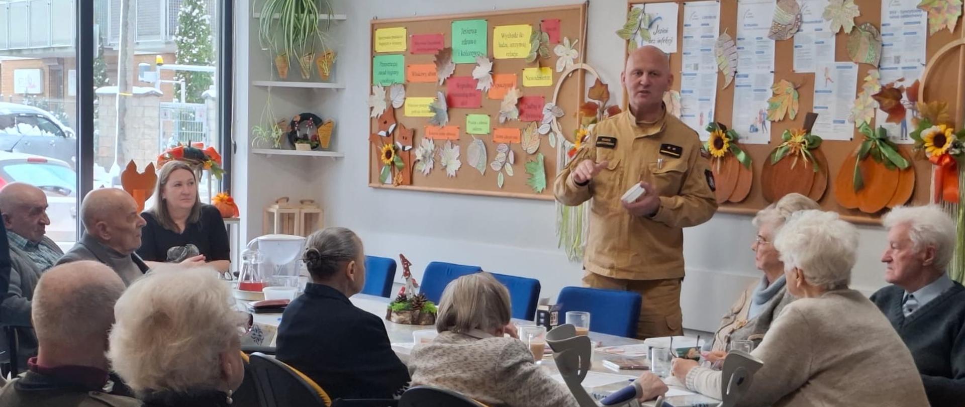 Ujęcie z sali, gdzie mężczyzna w mundurze strażackim (Marek Grodziński) przemawia do grupy starszych osób siedzących przy stole. Prezentuje trzymany w dłoniach mały biały przedmiot (prawdopodobnie czujnik). Wokół stołu siedzi około dziesięciu osób, głównie starsze kobiety, z których niektóre opierają się na laskach lub balkoniku. W tle, na ścianie, widoczna jest ta sama jesienna tablica informacyjna.