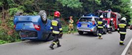 Samochód osobowy na dachu a obok strażacy i samochody służb.