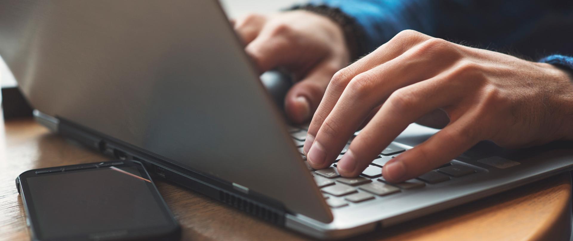 Dłonie na klawiaturze laptopa, telefon komórkowy.