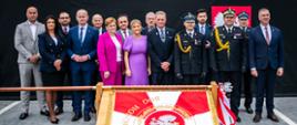 Na pierwszym zdjęciu widać grupę około trzynastu osób ustawionych do oficjalnej fotografii. Stoją na zewnątrz, na tle ciemnej sceny lub kurtyny. W centrum, na drewnianym stojaku, znajduje się czerwono-biały sztandar z polskim orłem i złotymi zdobieniami.
Osoby są ubrane bardzo elegancko. Część ma garnitury w granatowych i szarych kolorach, a kilka osób nosi mundury strażackie z czapkami i złotymi sznurami ceremonialnymi. Dwie kobiety stojące pośrodku wyróżniają się jasnymi ubraniami — jedna ma intensywnie różowy garnitur, druga lawendową suknię. Wszyscy patrzą w stronę aparatu i stoją w spokojnych, oficjalnych pozach.
Atmosfera zdjęcia jest uroczysta i reprezentacyjna.