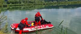 Kolorowa fotografia wykonana w pogodny dzień na akwenie. Na pierwszym planie dwóch strażaków ubranych w specjalne ubrania do pracy w wodzie. Jeden z nich znajduje się przy brzegu a drugi w pontonie. W środku pontonu znajduje się sprzęt ratowniczy.