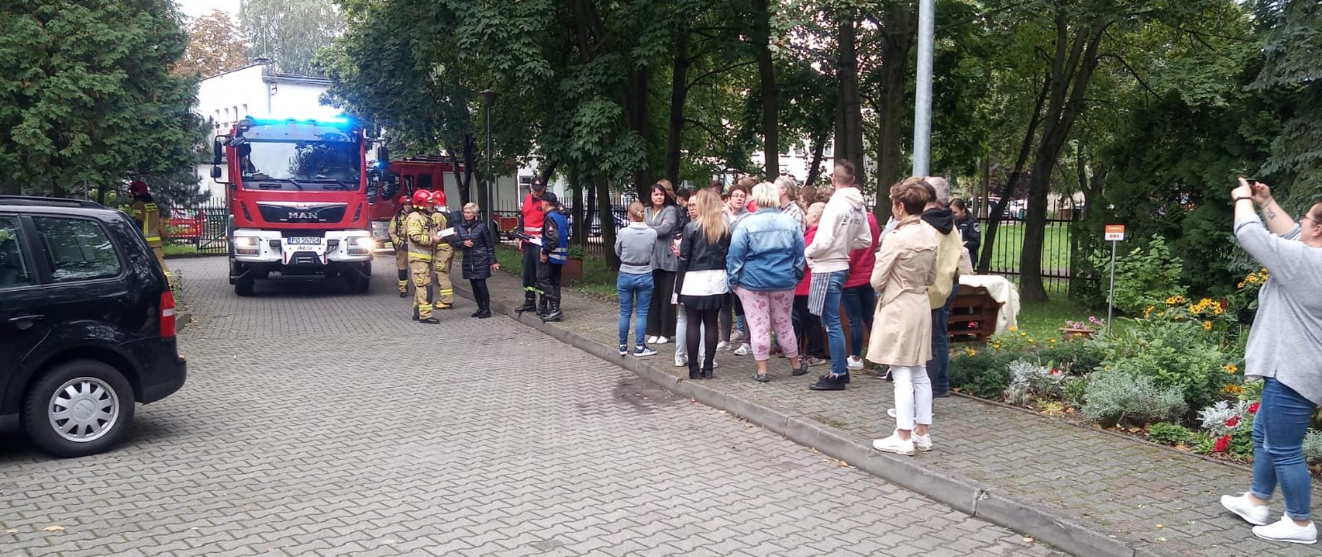 Osoby cywilne i strażacy stoją na drodze obok pojazdu strażackiego.