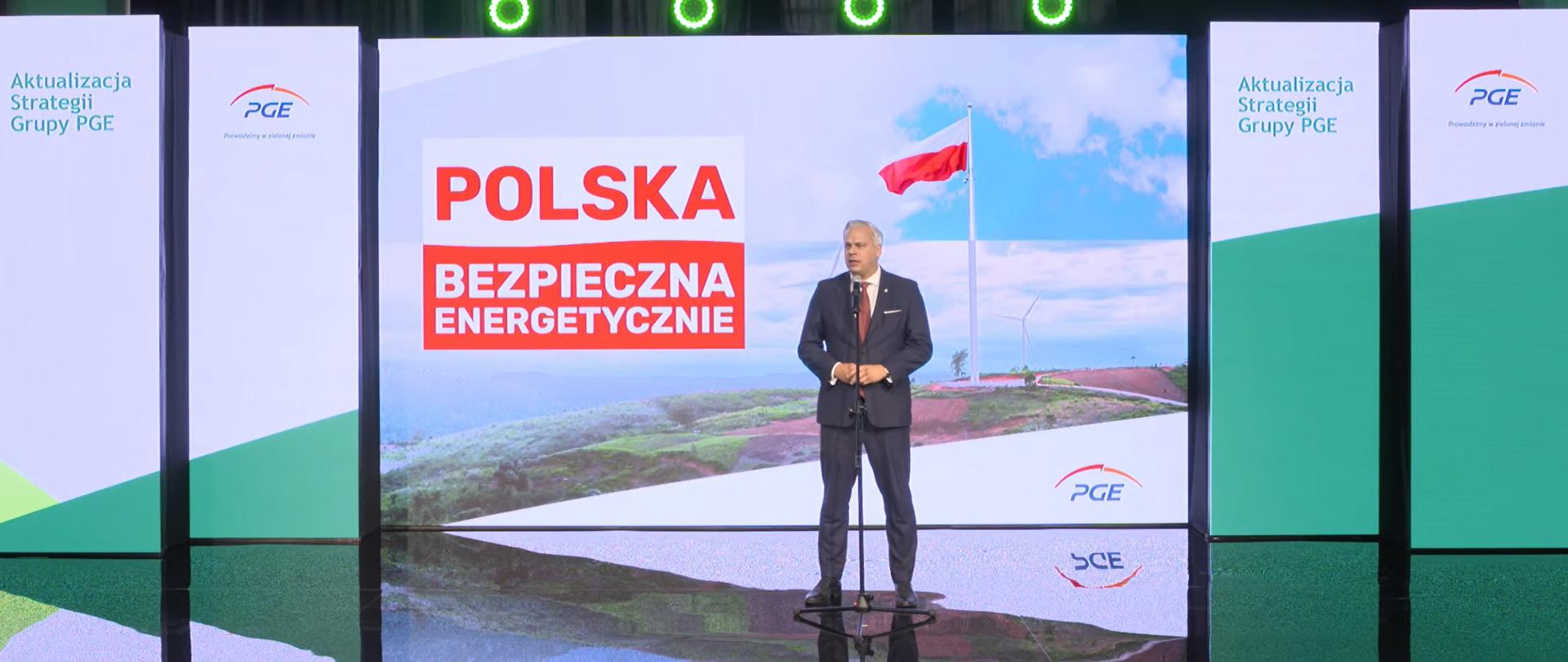 Wiceminister Karol Rabenda stoi za mikrofonem. W tle ścianka z napisem Polska bezpieczna energetycznie. 