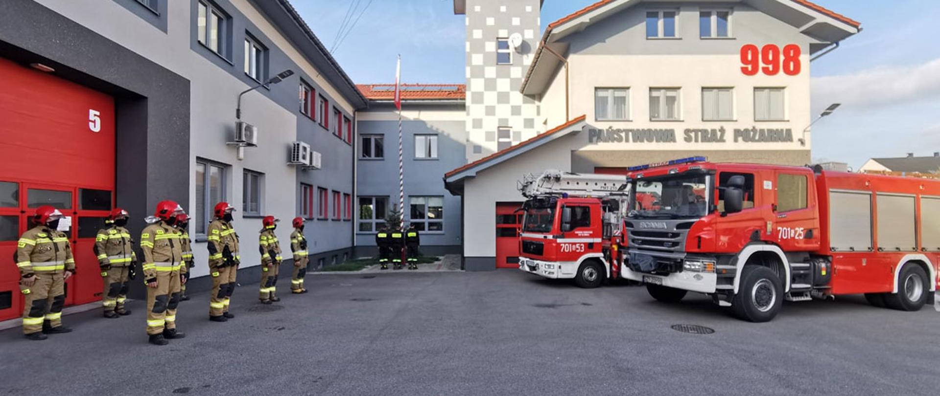Na zdjęciu jest dziedziniec główny Komendy Powiatowej Państwowej Straży Pożarnej w Żywcu. Z okazji Dnia Niepodległości strażacy śpiewają hymn, na maszt wciągana jest flaga narodowa. Na tle budynku ustawione są samochody pożarnicze, gaśniczy i podnośnik.