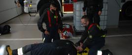 Na zdjęciu dwóch strażaków kursantów udziela pierwszej pomocy leżącemu na desce ortopedycznej pozorantowi pod nadzorem instruktora. W tle widzimy samochód pożarniczy ciężarowy.