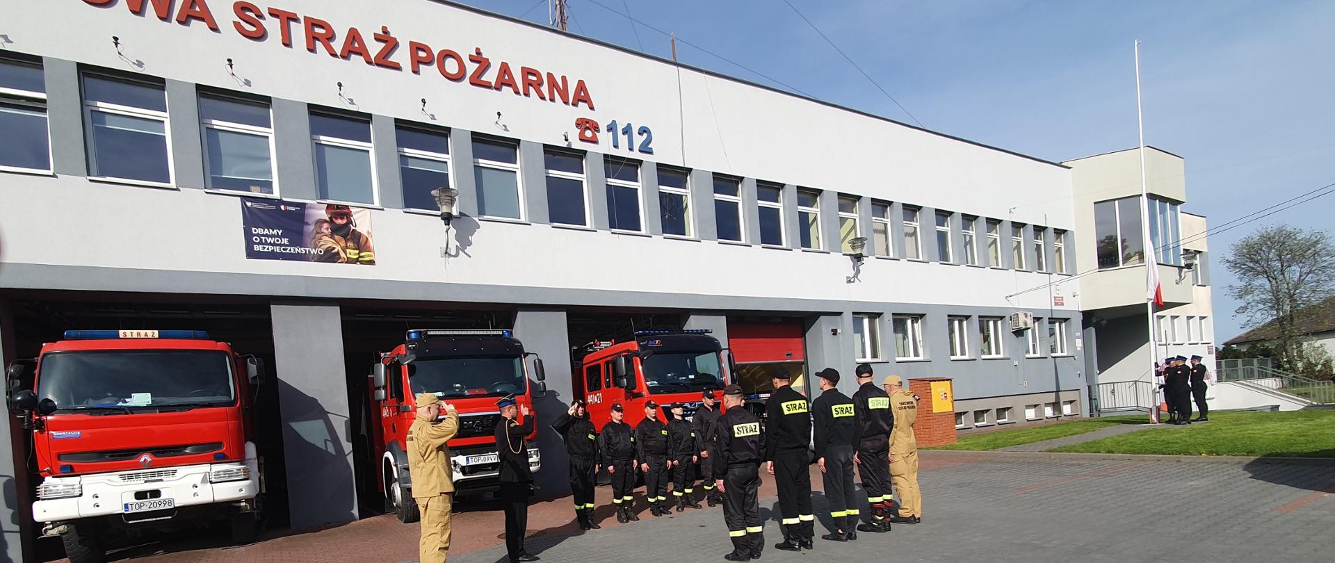 Zdjęcie przedstawia funkcjonariuszy stojących w szyku przed budynkiem KP PSP w Opatowie. Funkcjonariusze oddają honory podczas podnoszenia flagi państwowej.
