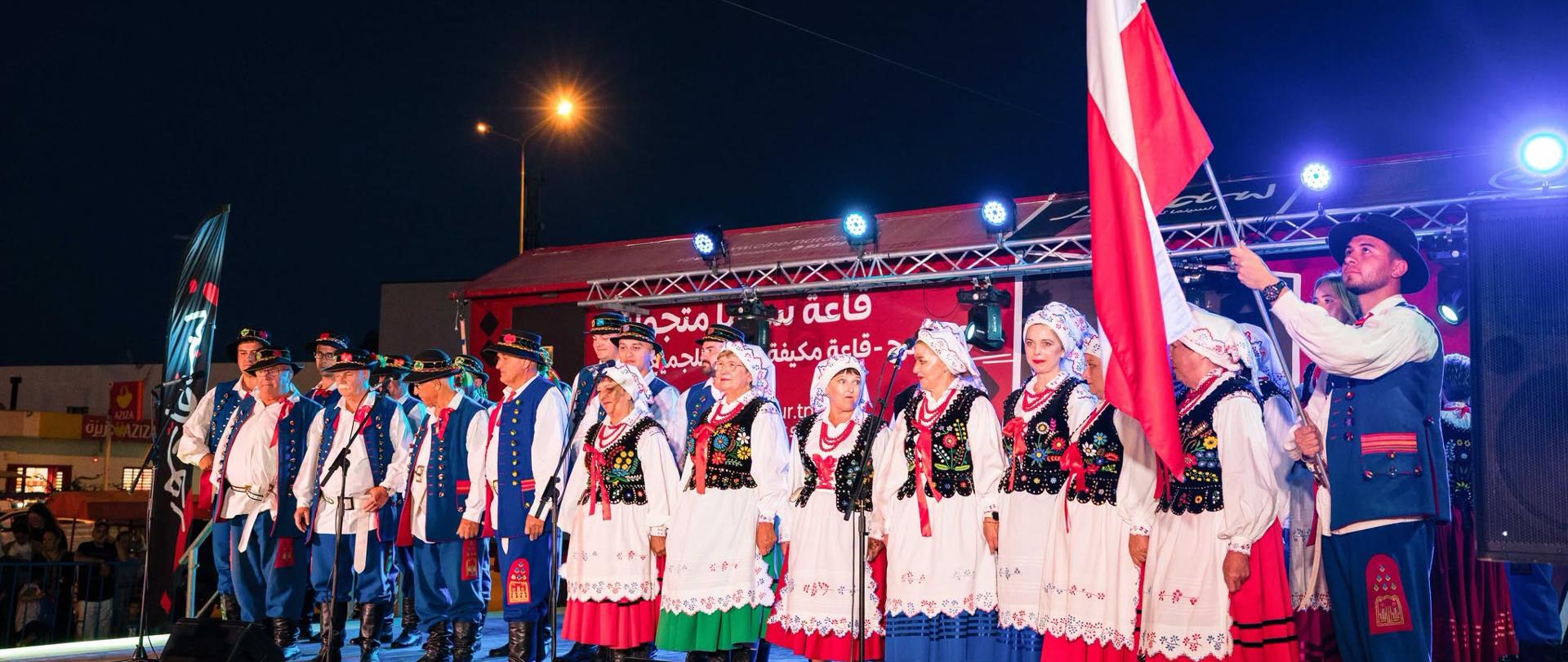 Występ polskiego zespołu folklorystycznego na scenie . Artyści w strojach ludowych stoją obok powiewającej biało-czerwonej flagi.