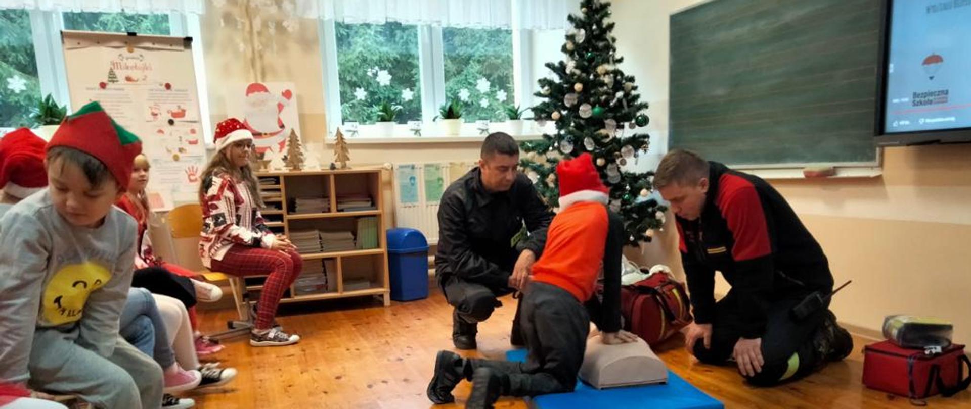 Zdjęcie przedstawia funkcjonariuszy Państwowej Straży Pożarnej w umundurowaniu służbowym, przed grupą uczniów szkoły podstawowej. Funkcjonariusze pochyleni nad fantomem tłumaczą zasady udzielania pierwszej pomocy.