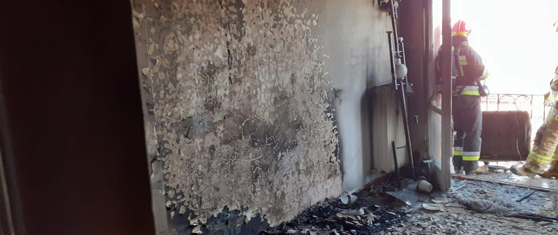 Spalony pokój z którego wyrzucono wyposażenie. W drzwiach balkonowych stoi strażak.