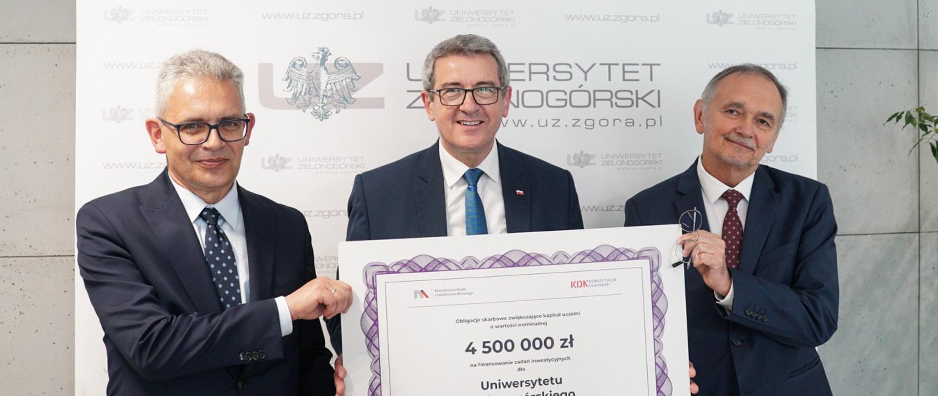 Minister i przedstawiciele uniwersytetu stoją prezentując obligacje.