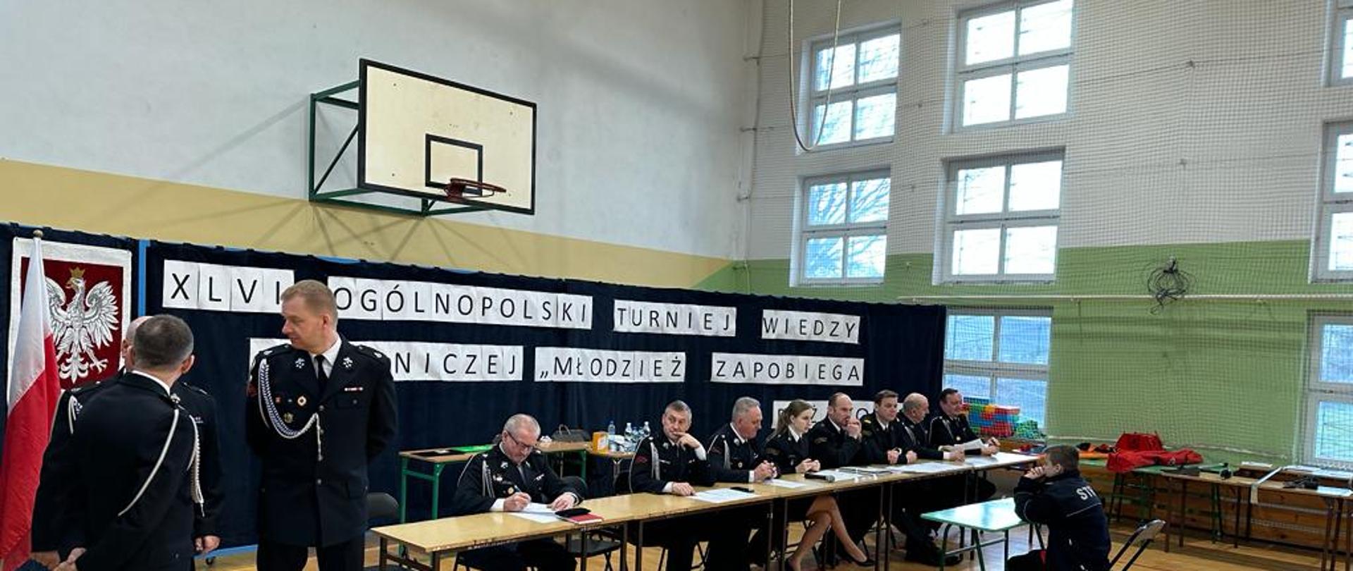 Zdjęcie wewnątrz pomieszczenia. Sala gimnastyczna. Widać osoby. Podłoga drewniana. Widać stoły oraz krzesła.