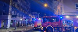 Zdjęcie wykonane w porze nocnej na miejskiej ulicy. Na ulicy samochody służb ratowniczych. Posiadają sygnalizację ostrzegawczą. W tle kamienica. W kadrze ujęta również latarnia oświetlenia ulicznego.